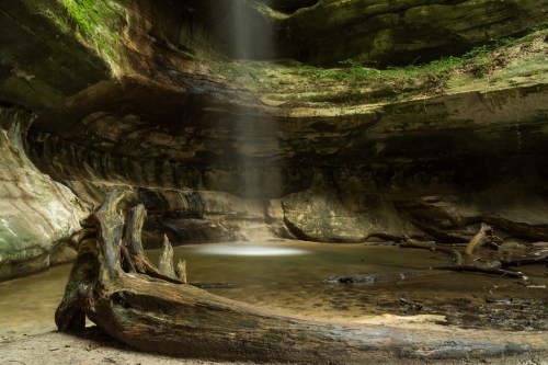 A serene cave with a small pool of water, illuminated by a beam of light filtering through the rocky ceiling.