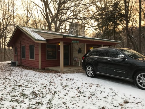 A cozy red cabin with a stone chimney, surrounded by snow and trees, with a black SUV parked in front.