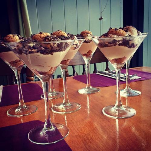 Five elegant dessert glasses filled with layered cream and topped with cookies, set on a wooden table.