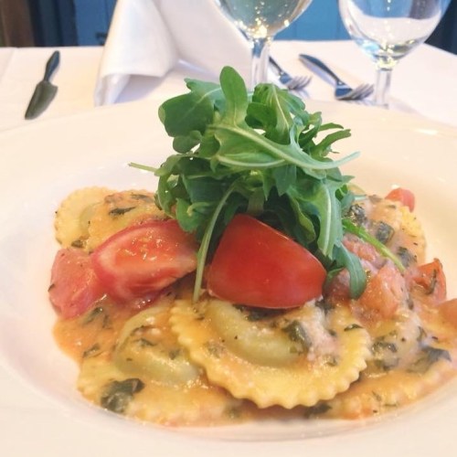 A plate of ravioli topped with fresh tomatoes and arugula in a creamy sauce, set on a restaurant table.