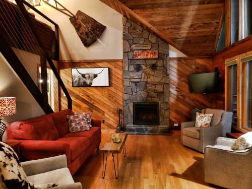 Cozy living room with a stone fireplace, wooden walls, red sofa, and large windows letting in natural light.