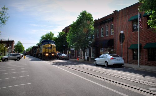 A train passes through a small town street lined with trees and brick buildings, with parked cars along the road.
