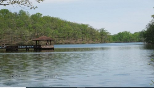 A serene lake scene with a wooden gazebo on a dock, surrounded by lush green trees and calm water.