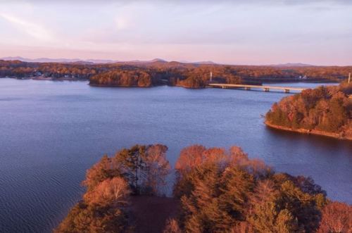 A serene lake at sunset, surrounded by trees and distant mountains, with a bridge crossing the water.