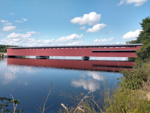 A red covered bridge spans a calm river, reflecting clouds and greenery under a blue sky.