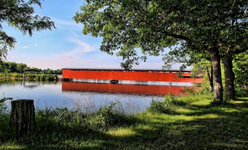 A red covered bridge spans a calm river, surrounded by lush greenery and trees under a clear blue sky.
