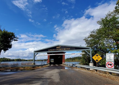 A historic covered bridge from 1887 spans a river, with a weight limit sign and cloudy sky in the background.