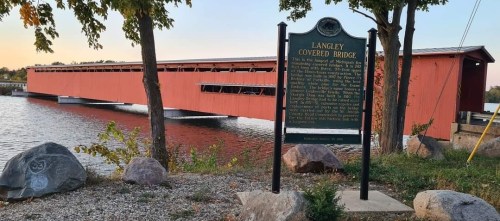 A red covered bridge spans a calm body of water, with a historical sign nearby and trees lining the shore.