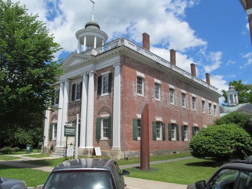 Historic brick building with white columns, surrounded by greenery and under a blue sky with clouds.