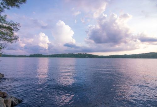 A serene lake scene at sunset, with calm waters reflecting fluffy clouds and a distant tree-lined shore.