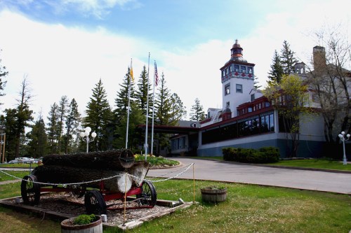 Historic building with a tower, surrounded by trees, featuring a wagon with logs in the foreground and flags flying.
