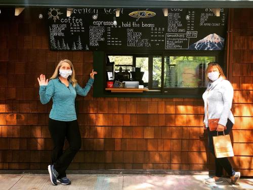 Two women in masks stand outside a coffee shop, pointing at the menu board. One holds a shopping bag.
