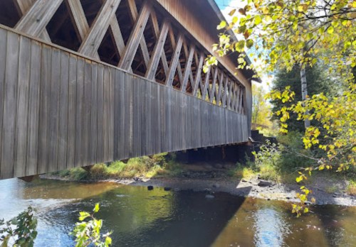 A wooden covered bridge spans a calm river, surrounded by trees with autumn leaves. Sunlight filters through the structure.