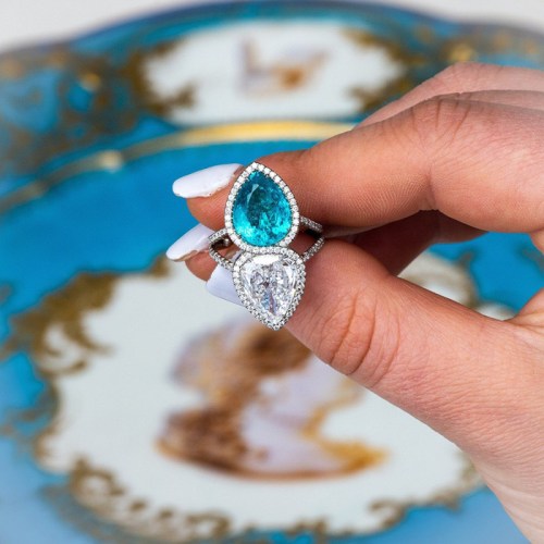 A hand holding a stunning ring featuring a teardrop-shaped turquoise stone and a pear-shaped diamond, set against a decorative background.