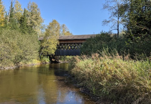 A covered bridge spans a calm river, surrounded by trees and tall grass under a clear blue sky.