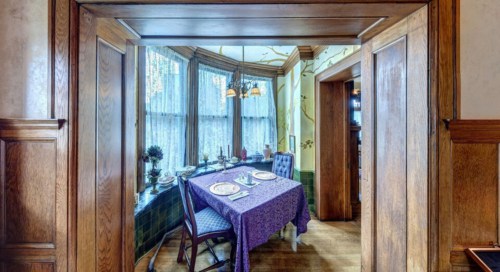 A cozy dining area with a purple tablecloth, set for two, surrounded by large windows and wooden accents.