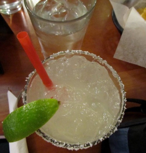 A close-up of a margarita with a lime wedge and a straw, next to a glass of water on a wooden table.