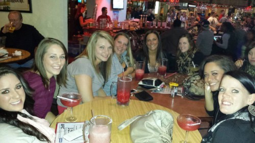 A group of smiling women sitting at a bar with drinks, enjoying a lively social atmosphere.