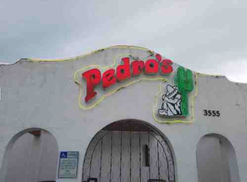 Sign for "Pedro's" featuring a cactus and a character, located on a white building with a cloudy sky above.