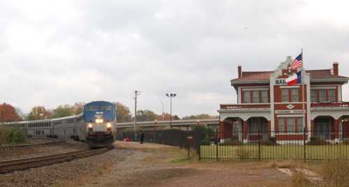 A train approaches a historic train station with a Texas flag, surrounded by trees and a cloudy sky.