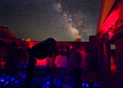 A starry night sky with the Milky Way, featuring a telescope and silhouettes of people in a dimly lit observatory.