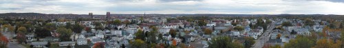 A panoramic view of a town with colorful autumn foliage, featuring buildings and distant hills under a cloudy sky.