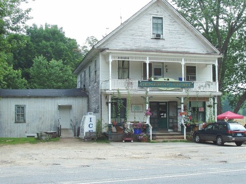A rustic general store with a porch, surrounded by greenery, and a parked car in front.