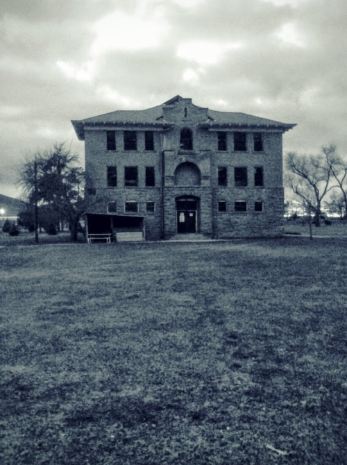 A large, old stone building with multiple windows, set against a cloudy sky and grassy foreground.