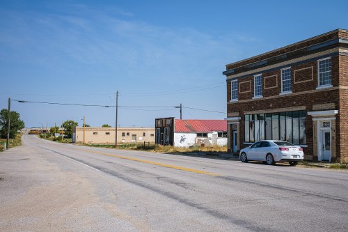 A quiet street scene featuring a brick building, a white car, and a few scattered structures under a clear blue sky.