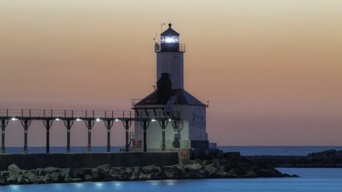 A lighthouse stands on a pier at dusk, with calm waters and a colorful sky in the background.