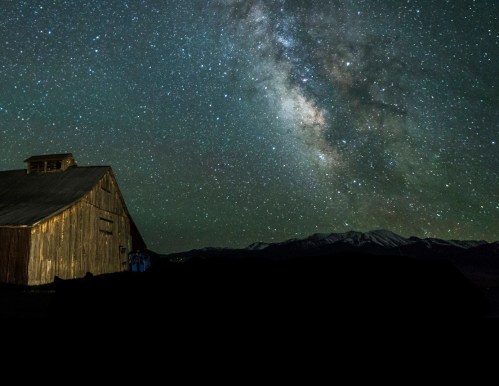 A rustic barn silhouetted against a starry night sky, featuring the Milky Way and distant mountains.
