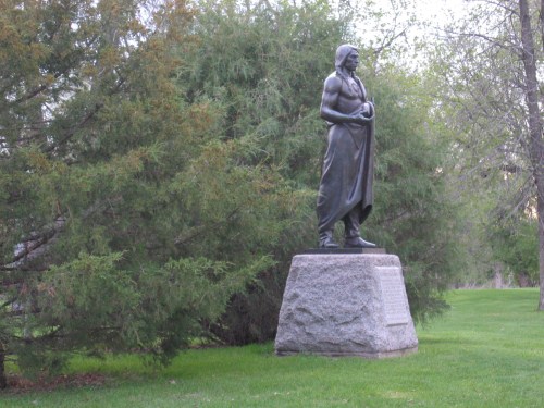 A bronze statue of a muscular figure draped in a robe, standing on a stone pedestal amidst green trees.