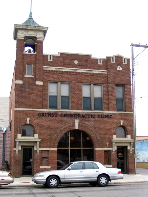 Historic brick building with a bell tower, featuring "Grunst Chiropractic Clinic" signage and a parked car in front.