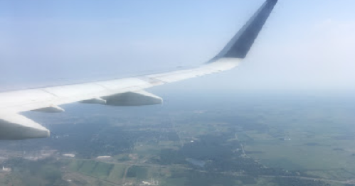 View from an airplane window showing a wing and a landscape of fields and distant hills below.