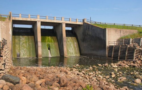 A concrete dam with water flowing over it, surrounded by rocks and grass under a clear blue sky.