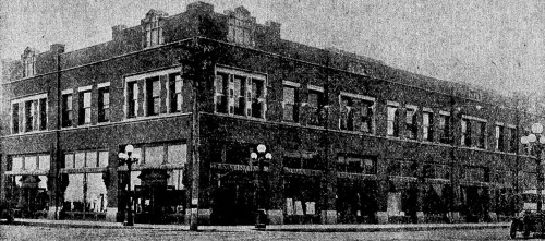Historic brick building with multiple windows and storefronts, featuring street lamps and pedestrians outside.