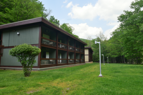 A two-story building with balconies, surrounded by green grass and trees under a partly cloudy sky.