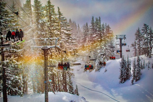 Skiers on a chairlift surrounded by snow-covered trees and a rainbow in a winter landscape.