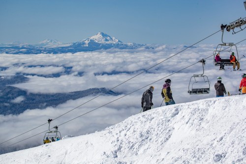 Skiers on a snowy slope with a chairlift, overlooking clouds and mountains in the background.