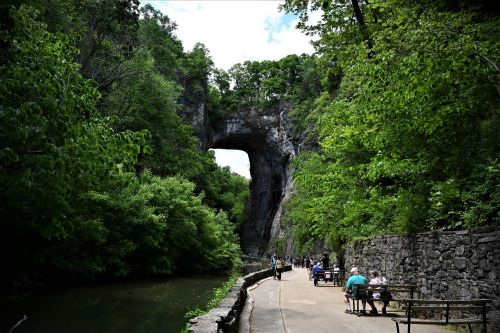 A scenic pathway alongside a river, featuring a large stone arch and lush greenery. People are enjoying the outdoors.