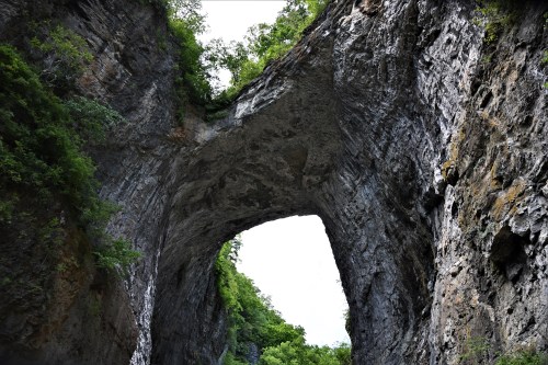 A natural stone arch surrounded by lush greenery and rocky cliffs under a bright sky.
