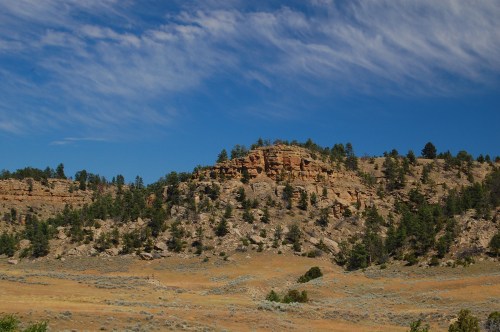 A rocky hillside with scattered trees under a blue sky with wispy clouds, set in a dry, open landscape.