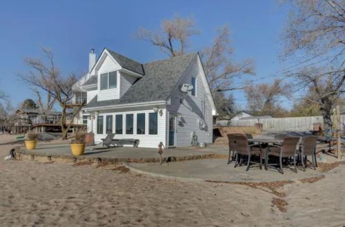 A cozy beach house with a patio and outdoor dining area, surrounded by sand and trees under a clear blue sky.