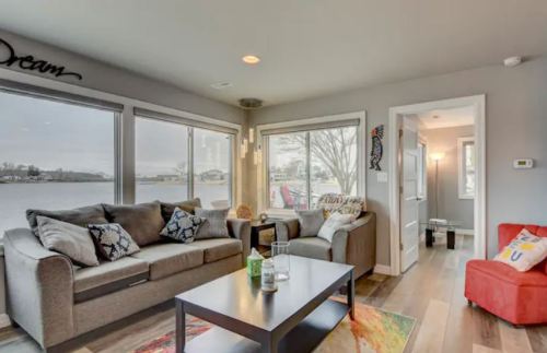 Cozy living room with large windows overlooking a lake, featuring a gray sofa, coffee table, and colorful decor.