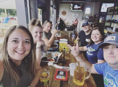 A group of six friends smiling and posing for a selfie at a bar, with drinks and food on the table.