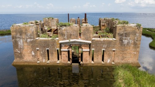 Aerial view of a weathered brick ruin surrounded by water and overgrown vegetation, reflecting a historical structure.
