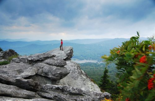 A person stands on a rocky outcrop overlooking a lush valley and distant mountains under a cloudy sky.