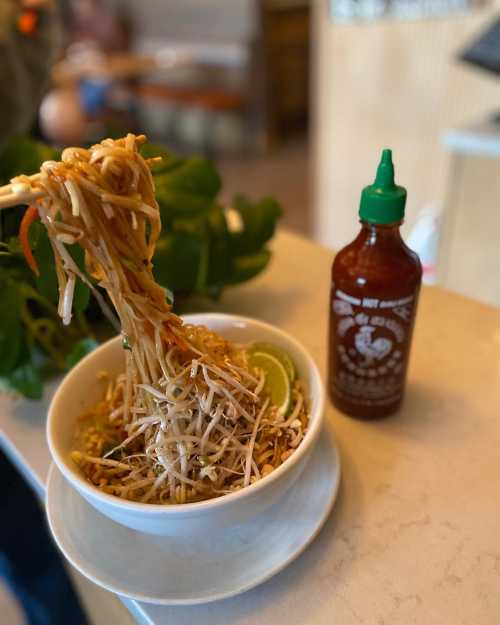 A bowl of pad thai with bean sprouts and lime, accompanied by a bottle of sriracha sauce on a table.