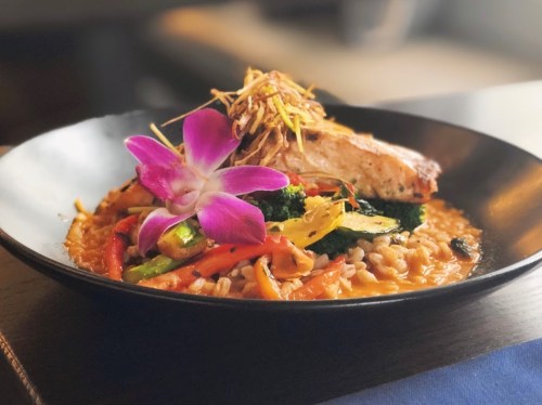 A beautifully plated dish featuring salmon, colorful vegetables, and a purple orchid garnish on a black plate.