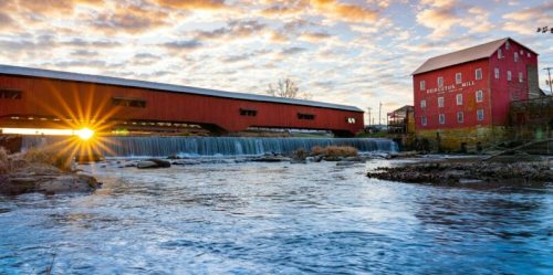 A red covered bridge spans a river, with a historic building nearby and a sunset casting a warm glow over the scene.